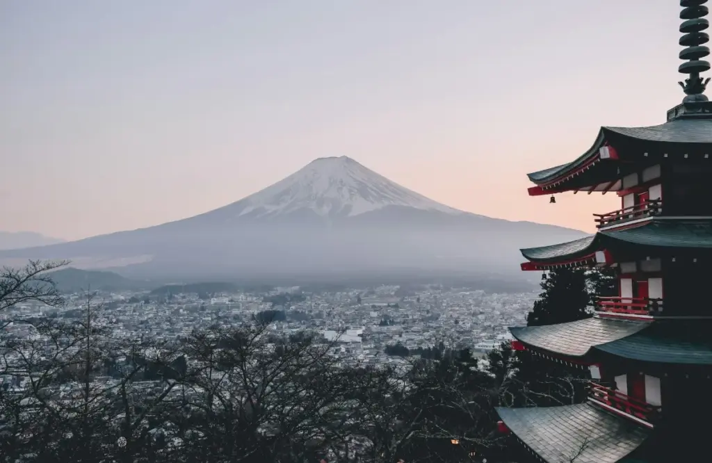 朝焼けの空を背景に、雪に覆われた富士山が見える風景。右手前には赤と黒の装飾的な多層塔が立ち、その下に広がる日本の都市全体が見える眺め