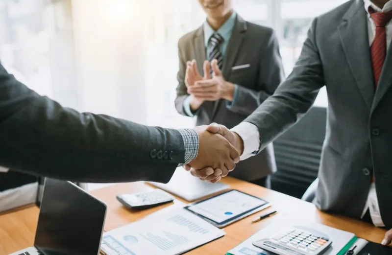 スーツを着たビジネスパーソン同士がオフィスで握手を交わし、背景では別の人物が拍手している商談成立の場面。