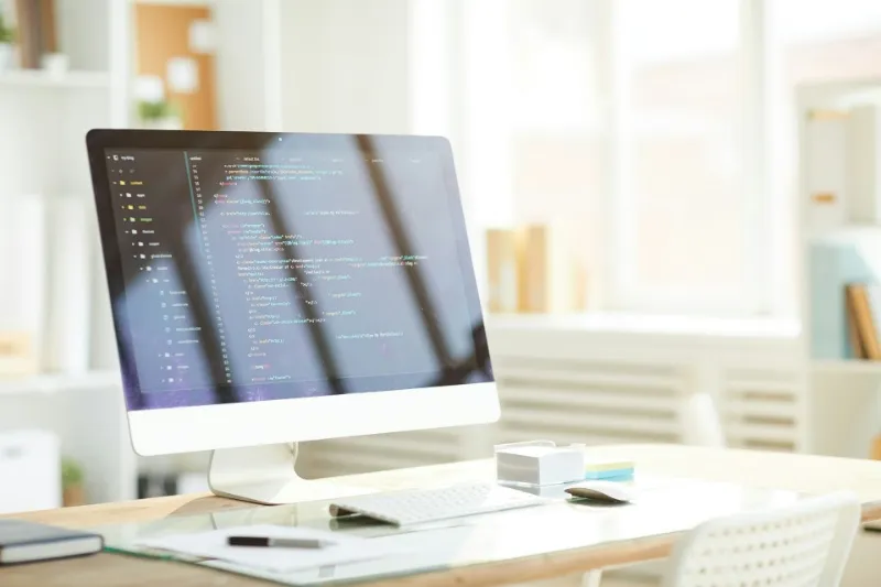 明るいオフィスのデスク上に置かれた大型ディスプレイにHTMLコードが表示されており、清潔で整った開発環境が写っている。