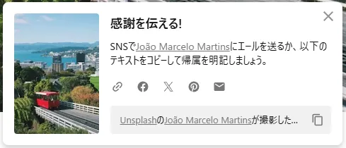 日本語の感謝メッセージ共有ダイアログ。左側に海岸線都市と鉄道の風景写真、右側に「感謝を伝える！」というタイトルと SNS 共有オプション（Facebook、X、Pinterest、メール）。下部に Unsplash のフォトクレジット「João Marcelo Martins が撮影した」と記載、閉じるボタンと画像 URL コピー機能がある